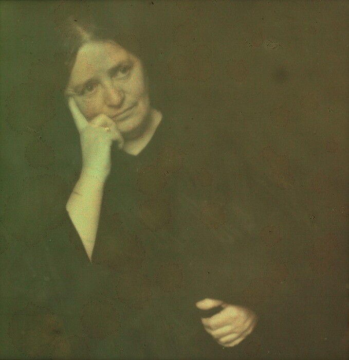 The image displays a woman from the waist up, seated with her head resting on her hand. She has long, dark hair, parted down the middle, falling past her shoulders. The woman's facial features are soft and her expression is neutral. She wears a dark garment covering her shoulders and a ring on one of her fingers. The background is a muted shade with a textured appearance.