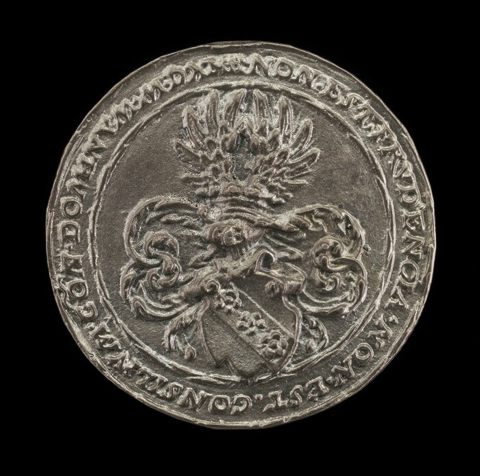 This is a photograph of a silver coin-like object with intricate designs. The coin features an elaborate coat of arms in the center, with detailed scrollwork and a helmet with a decorative plume above it. The outer edge of the coin is inscribed with text in an old script. The surface of the coin has a slightly worn texture, giving it an antiquated appearance. The photograph captures the intricacy of the carvings and the reflective metallic sheen of the coin against a plain black background.