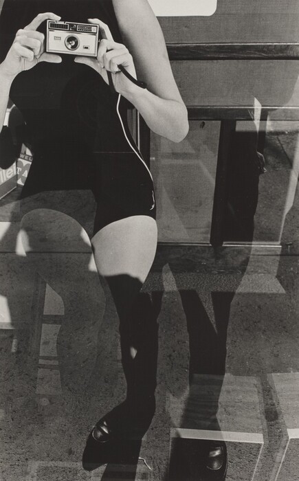 This is a photograph of a person holding a camera. The image captures an individual from the neck down, predominantly focusing on the act of holding a vintage box camera in a defined pose. The legs are prominently displayed against a reflective or transparent surface that creates multiple layers or shadows. The setting appears to be urban, suggested by the hard surfaces and linear shapes in the background. The overall tone is monochromatic, heightening the interplay between shadows and light.