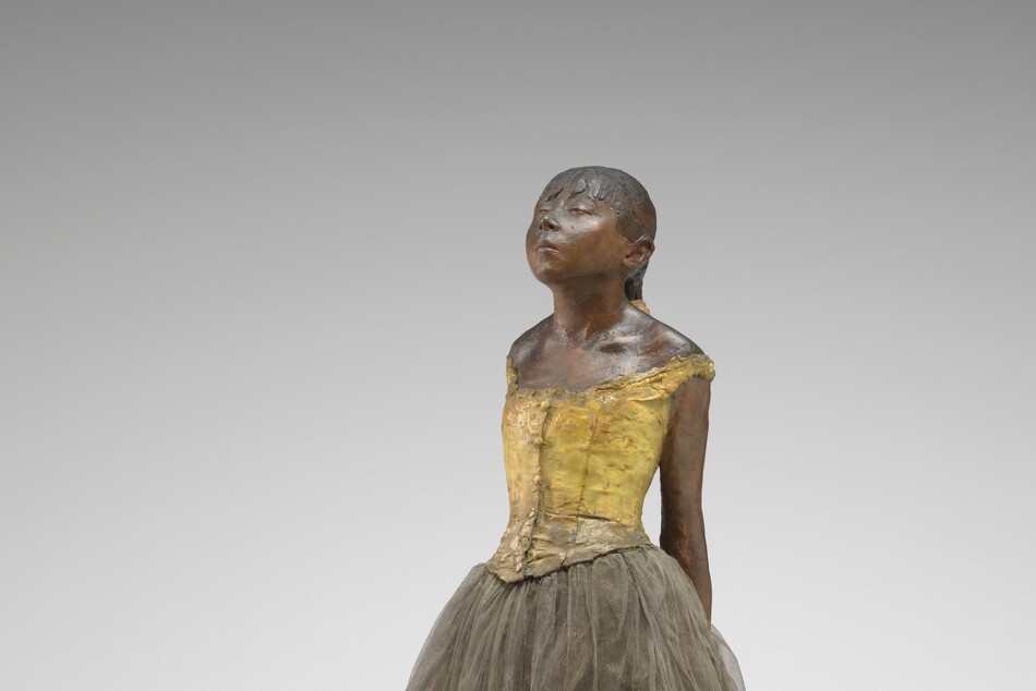 Sculpted with rich brown wax, a young ballerina stands with her arms straight, hands clasped behind her back, and one foot in front of the other on a square wooden base. Her body is angled to our left in this photograph. Both feet are splayed outward, and her right foot is placed far in front of her left. Bangs cover her forehead, and she has a heart-shaped, upturned face with a squat nose and slightly pursed lips. Her heavy-lidded eyes are nearly closed and her hair is pulled back and tied with a wide, cream-white ribbon. She wears a fabric costume with a sleeveless, gold-colored bodice, a gray tulle skirt, and ballet slippers. Her body is sculpted from dark brown wax, and a layer of wax covers her hair, bodice, and ballet slippers.
