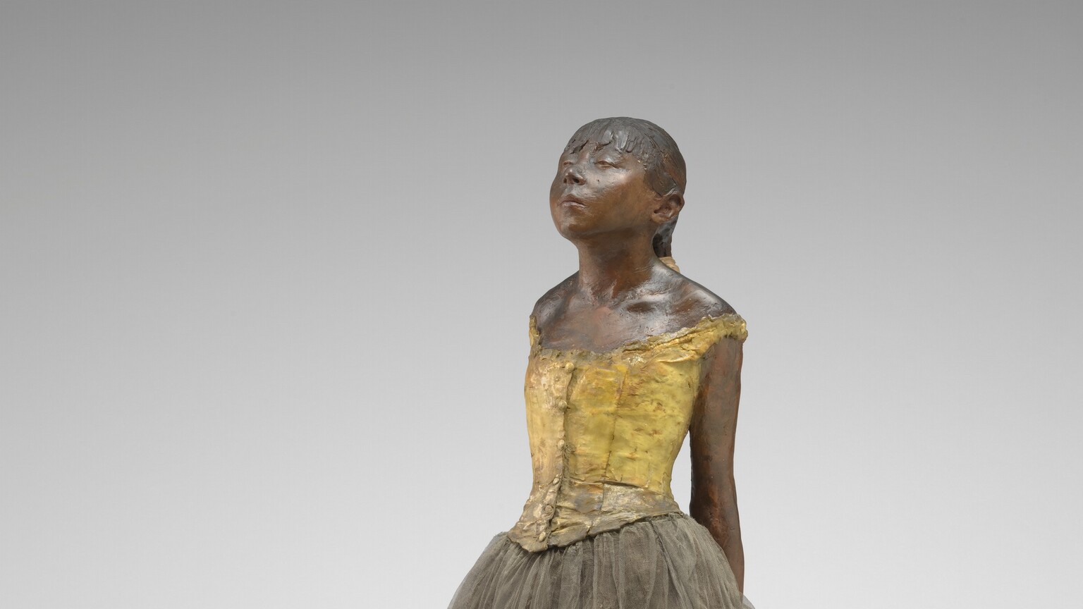 Sculpted with rich brown wax, a young ballerina stands with her arms straight, hands clasped behind her back, and one foot in front of the other on a square wooden base. Her body is angled to our left in this photograph. Both feet are splayed outward, and her right foot is placed far in front of her left. Bangs cover her forehead, and she has a heart-shaped, upturned face with a squat nose and slightly pursed lips. Her heavy-lidded eyes are nearly closed and her hair is pulled back and tied with a wide, cream-white ribbon. She wears a fabric costume with a sleeveless, gold-colored bodice, a gray tulle skirt, and ballet slippers. Her body is sculpted from dark brown wax, and a layer of wax covers her hair, bodice, and ballet slippers.