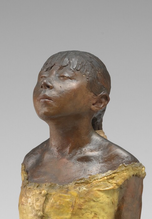 Sculpted with rich brown wax, a young ballerina stands with her arms straight, hands clasped behind her back, and one foot in front of the other on a square wooden base. Her body is angled to our left in this photograph. Both feet are splayed outward, and her right foot is placed far in front of her left. Bangs cover her forehead, and she has a heart-shaped, upturned face with a squat nose and slightly pursed lips. Her heavy-lidded eyes are nearly closed and her hair is pulled back and tied with a wide, cream-white ribbon. She wears a fabric costume with a sleeveless, gold-colored bodice, a gray tulle skirt, and ballet slippers. Her body is sculpted from dark brown wax, and a layer of wax covers her hair, bodice, and ballet slippers.