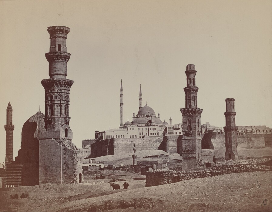 The image shows a historical architecture view. Multiple minarets rise into the sky, with a grand mosque complex featuring a large dome and two slender minarets in the distance. Several ancient, ornately carved minarets are visible in the foreground, with detailed stonework. The color palette is monochromatic with sepia tones. Stone buildings in the distance create a layered effect, leading towards the central mosque. The sky above is empty.