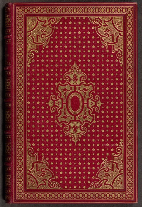 This is a photograph of a red book cover with gold embossed patterns. The central design features an intricate geometric motif enclosing an oval shape, surrounded by small gold stars or floral shapes on a red background. The corners have swirling gold motifs and the borders consist of symmetrical designs.