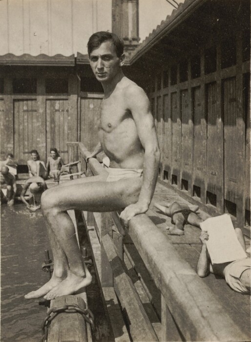 The image shows a man in a seated position on a wooden railing, which extends over a body of water, giving a partial view of his upper body, legs, and face. He is shirtless, wearing only a pair of swim trunks. His facial features include a narrow face with a straight nose, prominent cheekbones, and short dark hair that slightly falls over his forehead. There is no visible jewelry or handheld objects. Around him, several other individuals are seen, either standing in the water or lounging on the railing, one of whom is reading a piece of paper. The background features a wooden structure, possibly changing rooms or bathhouses, under a tiled roof with shadows cast on the walls. The scene is reminiscent of a swimming or bathing area, characterized by its casual, public atmosphere.
