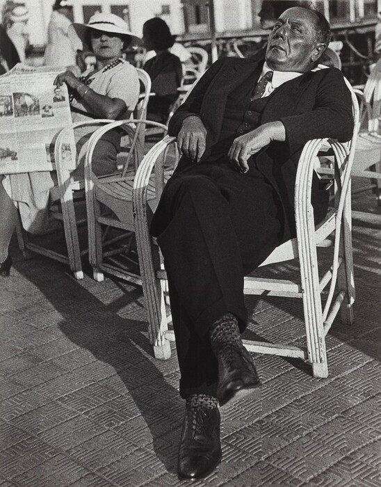 A man is sitting on a wicker chair in a leaning posture. He appears to be relaxing or enjoying the sunlight. The man has a round face with short dark hair, wearing a dark suit with a vest and tie, polished shoes, and patterned socks. Beside him, a woman is seated, wearing a white hat and holding a newspaper. Other people are partially visible in the background, seated on similar chairs. The setting seems to be outdoors, with some architectural structures in the blurred background.
