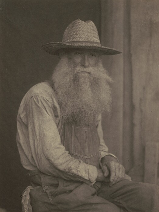 The image shows an older man sitting, with his torso visible from the waist up. He is seated facing slightly to the left, with his arms crossed casually. His facial features include a prominent, long, and full beard, and he has a large hat that casts a shadow over his eyes. His hair blends into the beard and appears to be as long and full as the beard itself. He is dressed in a light-colored, long-sleeved shirt underneath a pair of dungarees, with one visible pocket on the chest. The background is plain and subdued, with a textured surface behind him.