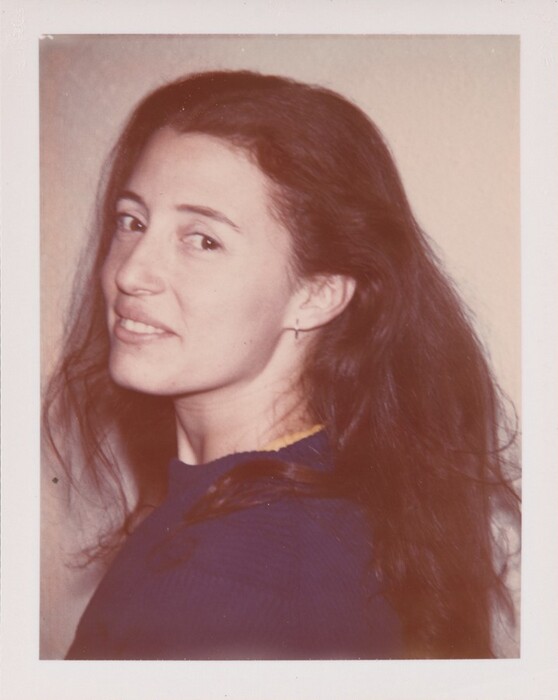 The image depicts a woman with dark brown hair in a blue ribbed top against a light-colored wall. The woman is looking slightly to the side with a subtle smile.
