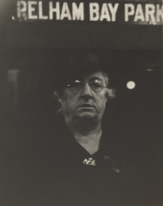 The image shows an older woman facing the camera with a solemn expression. She is wearing glasses, a hat, and a dark coat fastened with a brooch. There is a sign above her head that reads "PELHAM BAY PARK" in large uppercase letters. The background is dimly lit.