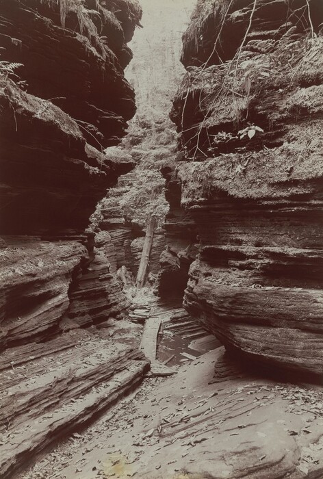 Two rocky, striated walls nearly fill this vertical black and white photograph. Through a narrow gap, like an open zipper, more striated rocks are visible.