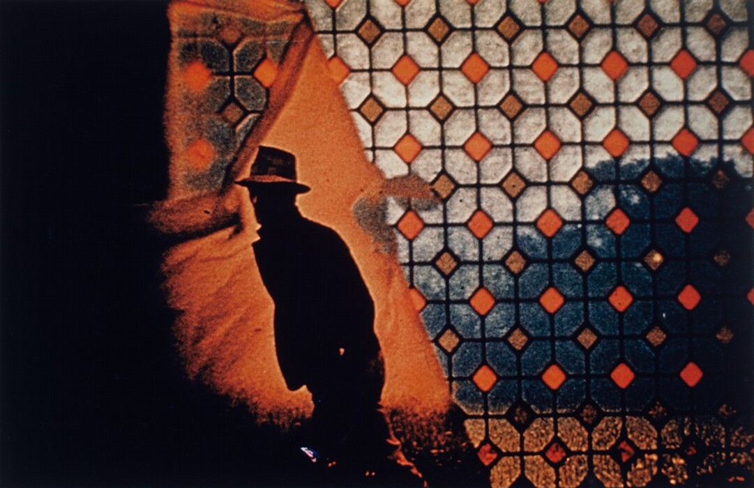 The image features a pattern of repeating octagonal shapes and diamond lines forming a grid with warm hues of orange and brown. In the foreground, an abstract silhouette of a man wearing a hat appears. The pattern of geometric shapes on the right side resembles a stained glass window, providing a backdrop to the outlined figure, creating a juxtaposition between the structured grid and the freeform silhouette.
