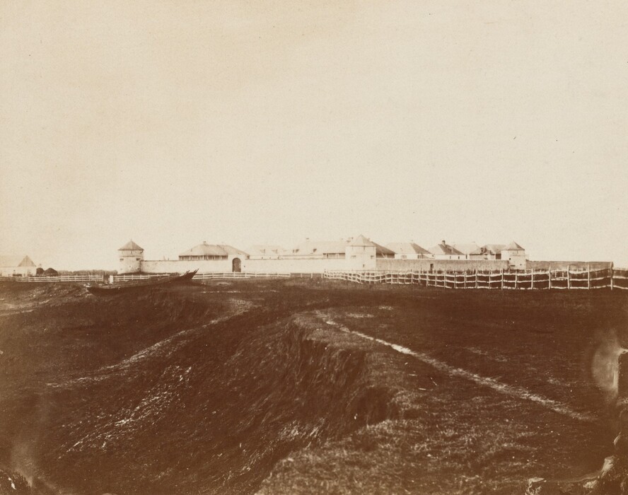 The image shows a view from a slightly elevated position looking across land towards a fort or settlement enclosed by a wooden fence. The horizon line is positioned about two-thirds up from the bottom of the image, primarily showing the structures of the fort with a few buildings visible beyond the fence. The image is in sepia tones, giving a vintage monochrome effect, emphasizing light and dark contrasts. The land in the foreground is uneven with trenches or ruts leading down to the fort in the distance.