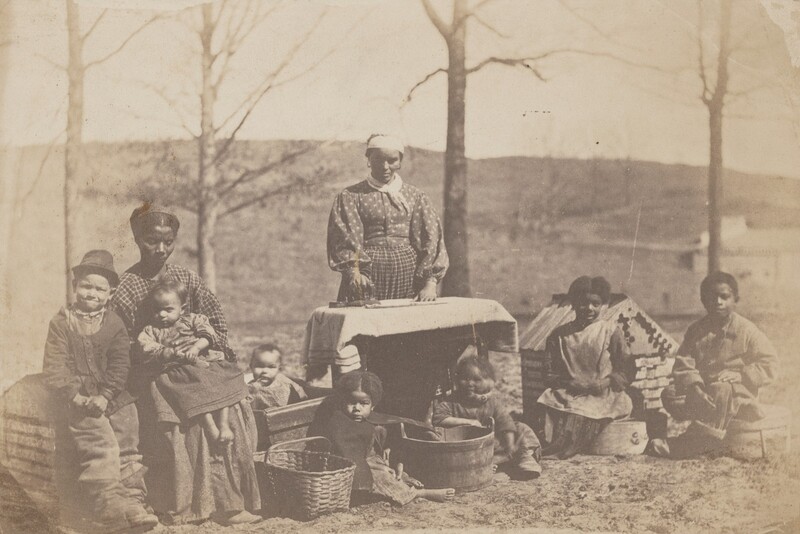 The image shows a group of individuals gathered outdoors around washing equipment. In the center, a woman stands with one hand on a table covered with a cloth. To the left, a seated woman has her arm around a small child. A young boy stands beside them. In front of the standing woman, another child sits next to a basket. On the right, three children sit on the ground, one inside a wooden tub. The background features bare trees, a hillside, and structures in the distance, suggesting a rural setting.