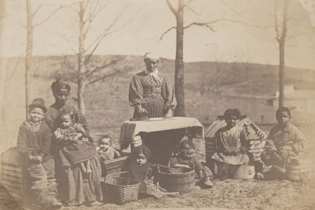 The image shows a group of individuals gathered outdoors around washing equipment. In the center, a woman stands with one hand on a table covered with a cloth. To the left, a seated woman has her arm around a small child. A young boy stands beside them. In front of the standing woman, another child sits next to a basket. On the right, three children sit on the ground, one inside a wooden tub. The background features bare trees, a hillside, and structures in the distance, suggesting a rural setting.