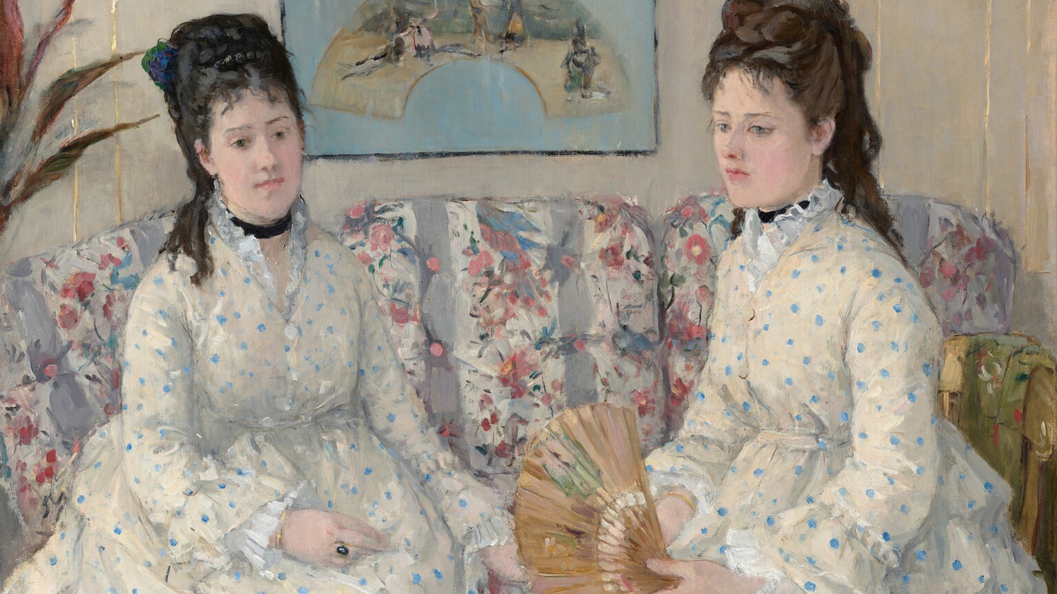 Shown from the lap up, two women with pale skin and dark hair pulled up and back, wearing white dresses with baby-blue polka dots, sit on a couch in this horizontal portrait painting. Both women have straight, dark brows, delicate noses, smooth skin, and their small, rose-pink mouths are closed. Wispy bangs brush their foreheads, and their hair is piled high with long ringlets falling down their backs. Both wear black ribbons like chokers around their necks. Their dresses have high necks with ruffles along the necklines, ruffles at the wrists, and are gathered under the bust. They sit angled in toward each other. The woman on our left has black hair, and she looks down and to our right with dark eyes. She wears a gold ring with a dark, oval stone on one hand resting in her lap. The other woman has chestnut-brown hair and looks down and off to our left with ice-blue eyes. She holds an open fan in her hands in her lap. The fabric on the couch has vertical white and lilac-purple stripes, and is overlaid with a pattern of pink flowers and green leaves. A framed picture hanging on the bone-white wall behind the women shows an arched painting against a sky-blue background. A few spiky leaves from a houseplant are cut off by the left edge of the painting. The portrait is loosely painted throughout, especially in the couch and background. The artist signed the painting in the upper right corner, “Berthe Morisot.”