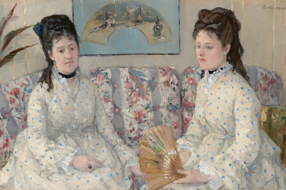 Shown from the lap up, two women with pale skin and dark hair pulled up and back, wearing white dresses with baby-blue polka dots, sit on a couch in this horizontal portrait painting. Both women have straight, dark brows, delicate noses, smooth skin, and their small, rose-pink mouths are closed. Wispy bangs brush their foreheads, and their hair is piled high with long ringlets falling down their backs. Both wear black ribbons like chokers around their necks. Their dresses have high necks with ruffles along the necklines, ruffles at the wrists, and are gathered under the bust. They sit angled in toward each other. The woman on our left has black hair, and she looks down and to our right with dark eyes. She wears a gold ring with a dark, oval stone on one hand resting in her lap. The other woman has chestnut-brown hair and looks down and off to our left with ice-blue eyes. She holds an open fan in her hands in her lap. The fabric on the couch has vertical white and lilac-purple stripes, and is overlaid with a pattern of pink flowers and green leaves. A framed picture hanging on the bone-white wall behind the women shows an arched painting against a sky-blue background. A few spiky leaves from a houseplant are cut off by the left edge of the painting. The portrait is loosely painted throughout, especially in the couch and background. The artist signed the painting in the upper right corner, “Berthe Morisot.”
