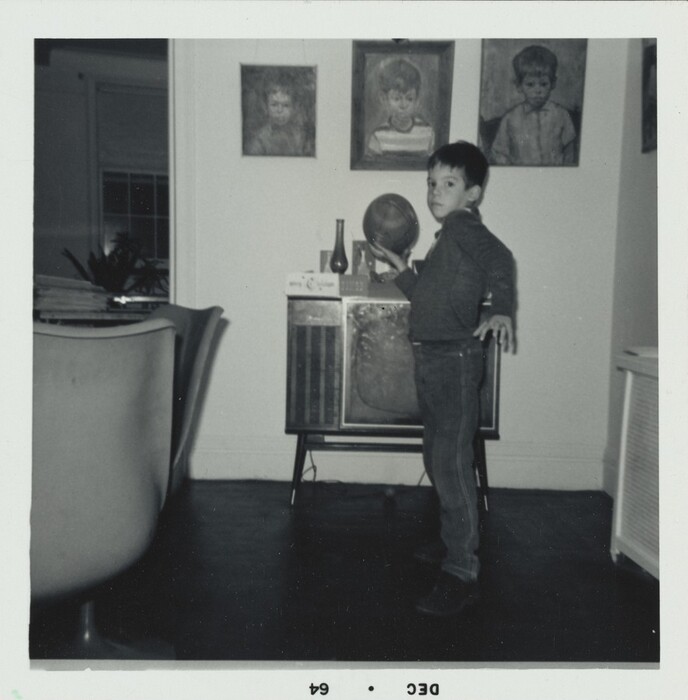 This is a photograph of an interior living space. It features a television set on a stand with a rectangular screen, glass bottle, and globe on top. Three framed portraits of children hang on the wall in the background. An armchair, stack of books or magazines, window with drapery, and plant complete the scene. The room has classic mid-century furnishings and decor.