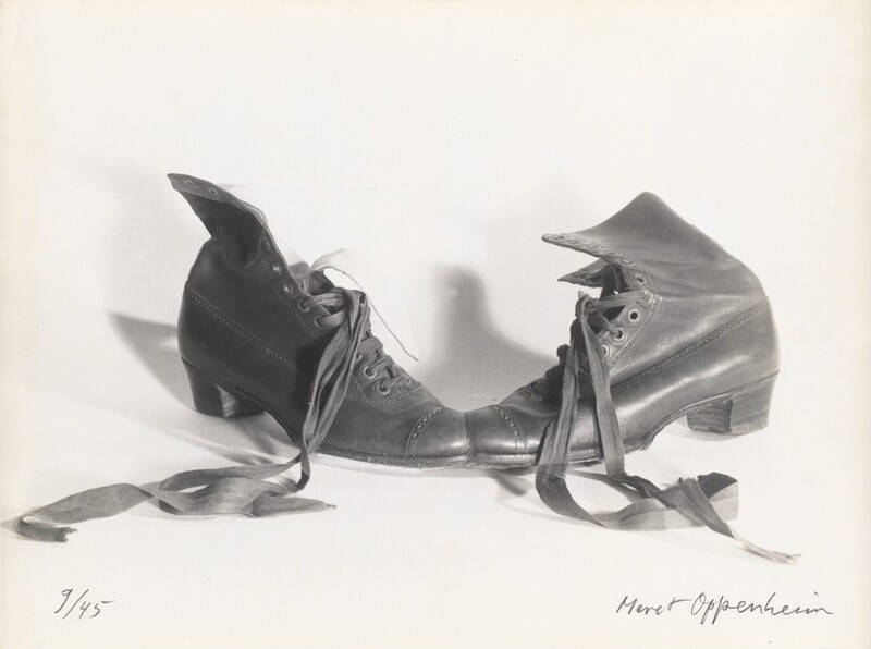 The toes of two leather lace-up boots face each other and, on closer inspection, are conjoined in this horizontal black and white photograph. The half-laced, ribbon-like laces of both boots are undone and splay out toward us almost symmetrically. The shaft of the left boot curls open, and the tongue lies close to its laces. The shaft of the right boot angles away from us. Both boots are decorated with patterned bands of punched holes over the toes, up the sides of the laces, and back along the upper, and both have square, stacked heels. The artist wrote “9/45” in the lower left corner and signed the lower right, “Meret Oppenheim,” both in graphite.