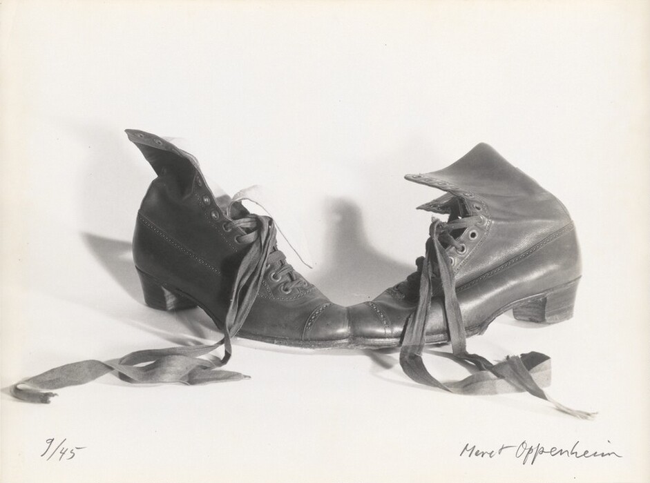 The toes of two leather lace-up boots face each other and, on closer inspection, are conjoined in this horizontal black and white photograph. The half-laced, ribbon-like laces of both boots are undone and splay out toward us almost symmetrically. The shaft of the left boot curls open, and the tongue lies close to its laces. The shaft of the right boot angles away from us. Both boots are decorated with patterned bands of punched holes over the toes, up the sides of the laces, and back along the upper, and both have square, stacked heels. The artist wrote “9/45” in the lower left corner and signed the lower right, “Meret Oppenheim,” both in graphite.