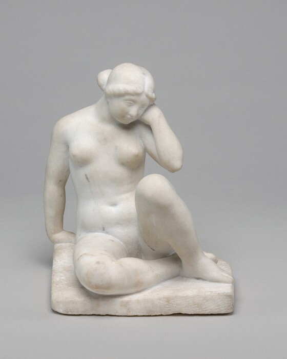 A nude woman sitting with bent knees is carved from white marble in this freestanding sculpture. In this view, the woman’s body faces us. Her right knee, to our left, is bent so that leg lies along the ground. Her feet are together, and the other bent leg is upright. She holds her left hand, to our right, to that cheek so the elbow is close to the raised knee. She leans onto her other hand, which is placed just behind her with the fingers facing in. Her round face tips toward her raised hand, and she has delicate features and wide cheeks. Her hair is parted down the middle, rolled along her hairline, and is pulled back. The square base on which she sits is carved from the same piece of marble. The sculpture is photographed against a gray background.