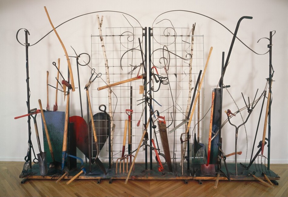 This sculpture is made up of about three dozen shovels, axes, pitchforks, hammers, scissors, saws, wrenches, and other tools arranged across a narrow wooden platform, which supports a free-standing, dark gray gate. The gate form is created by two black poles at the center and one on each narrow end to make two wide doors. Flat metal strips curve in long, shallow, horizontal S-shapes across the top of each door. Widely spaced wire grids extend from the central poles, about halfway across each door. Two thin trunks or branches of white birch trees stand behind the wire, one to each side. The handles of the tools create mostly vertical lines across the gates. The tools lie or sit on the platform, or lean against the gates. A few, including shears, hand drills, and calipers are affixed to the central posts or wire. A few objects stand out. At the leftmost edge of the platform, a propeller leans against the left-hand post. One shovel near the propeller and another near the right end of the platform have been cut across their blades, so they rest flat on the platform, as if thrust into dirt ground. A heart-shaped piece of sheet metal sits behind the gate, to our left. It comes just over halfway up the full-sized shovel in front of it, and is red on the left half and streaked with brown and black on the right half. The handles of a few of the tools project outward, toward us, including a pickaxe and pliers near the middle. The room in which this sculpture stands has a white wall and a wood floor, with the narrow planks laid diagonally to create radiating Vs.