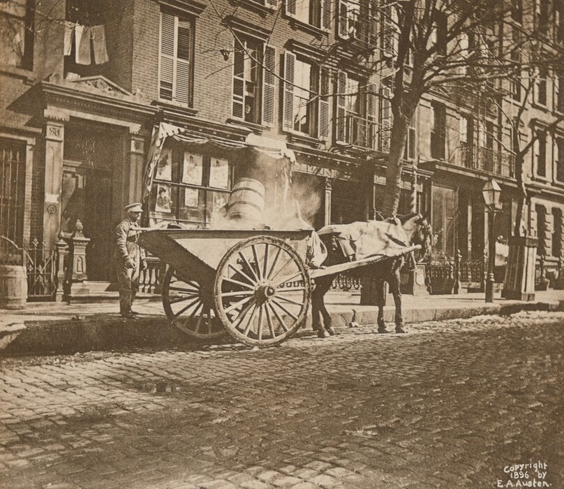 This is a photograph of a street scene with a horse-drawn cart. The image captures a cobblestone street lined with brick buildings. There is a horse-drawn cart with large wooden wheels standing by the curb, partially filled with material and a large barrel. The horse appears to be draped with fabric and is harnessed to the cart. The buildings have numerous windows, some with shutters, and several front entrances are visible, one of which has an ornate design. A lamppost and bare tree are also present.
