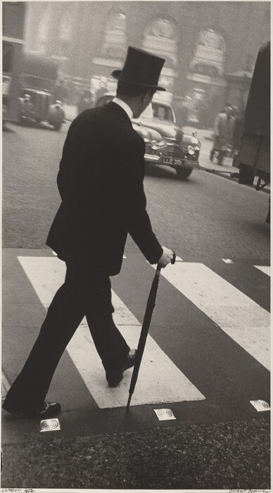 This is a photograph of a person crossing a street in a city. The individual is wearing formal attire and holding an umbrella while walking over a zebra crossing. In the background, there are classic cars and architectural buildings. The image is in black and white.
