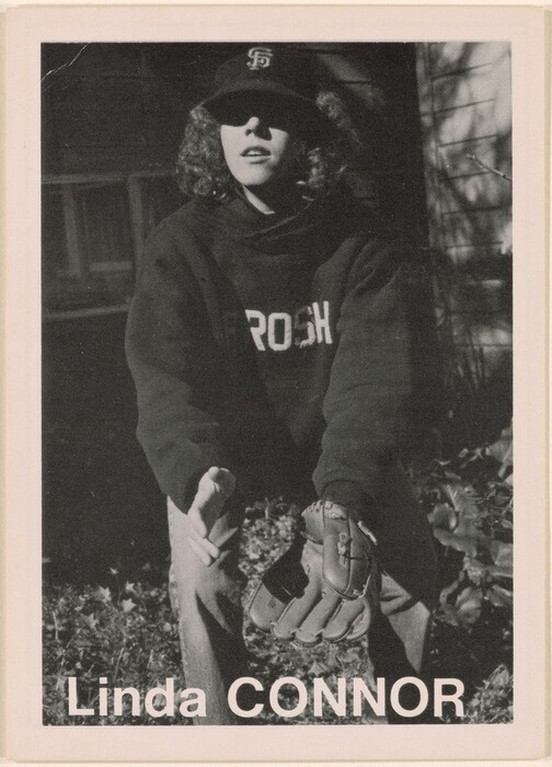 The image shows a person in a crouched position, with the upper body visible. They are positioned as if preparing to catch something with their arms reaching forward. The person has curly hair that extends below the edges of a dark baseball cap. Their facial features are partially shadowed, with a slightly open mouth. They wear a dark sweatshirt with partial text reading "ROSH" visible, and a baseball glove is on their left hand. The surrounding area includes foliage on the ground, with part of a building's side in the background, which is partially shadowed. The name "Linda CONNOR" is printed at the bottom of the image.
