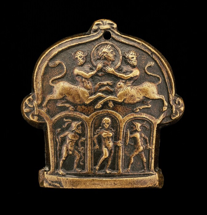 This is a photograph of a decorative metallic artifact. The object features intricate carvings depicting a mythological scene. The upper portion showcases two centaur-like creatures facing each other, each with a human torso and a feline lower body. Between them is a circular emblem containing the profile of a head. Below are arched niches containing three human figures stylized in dynamic poses. The figures carry various small adornments or instruments. The artifact exhibits detailed craftsmanship and a luxurious antique finish, possibly gilded, emphasized by the glowing gold-bronze hue against a darker background.