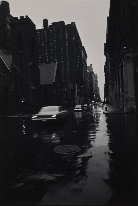 The image shows a wet city street at twilight or early evening. Car headlights move towards the viewer, creating a sense of motion. Tall, dark buildings obscure the horizon and lead away into the distance like a canyon. The sky is a bright, overcast glow above the buildings. The scene is monochromatic with deep blacks and grays dominating, and lighter tones reflecting in the wet road. The photograph captures a gritty, urban atmosphere with doorways and windows mostly in shadows, adding a moody quality. Beyond the intersection, dim and vague hints of city life can be seen, contributing to a sense of urban anonymity.