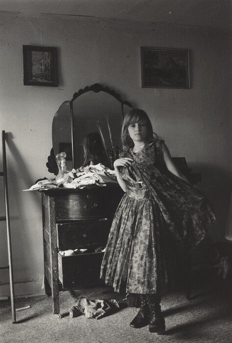 The image shows a young girl standing indoors. She is facing the camera with her head slightly turned to one side, her right hand holding part of her dress. She has a fair complexion and long, light-colored hair. The girl is wearing a patterned knee-length dress with a sleeveless design and dark lace-up boots. There is a dresser behind her with a large mirror and various items on it. Some drawers are slightly open. The walls have two framed paintings, and there is a piece of furniture to the left. The room is carpeted.