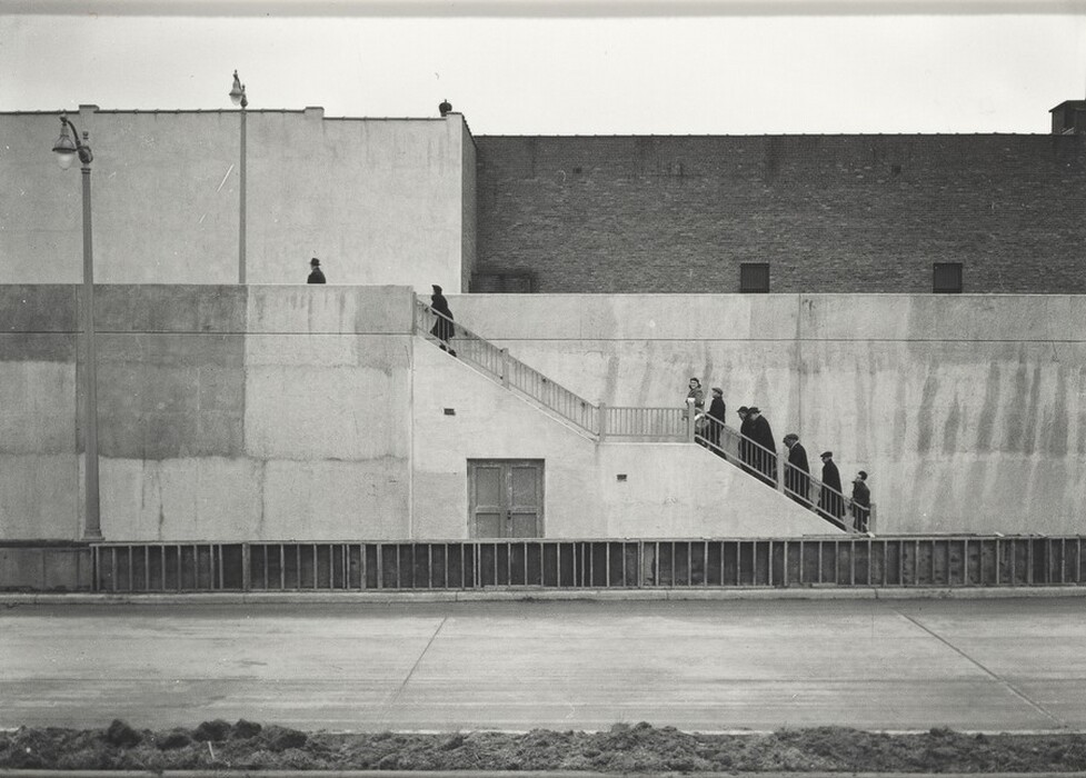 This is a photograph depicting multiple individuals climbing an outdoor staircase next to a large building. The image showcases urban architecture with a contrast between light concrete walls and the dark figures on the stairs. Two lamp posts are visible near the building, adding to the geometric composition. The scene conveys a sense of movement in an urban environment, emphasizing shapes and lines. The monochrome image accentuates the interplay of light and shadow, highlighting textures and forms.