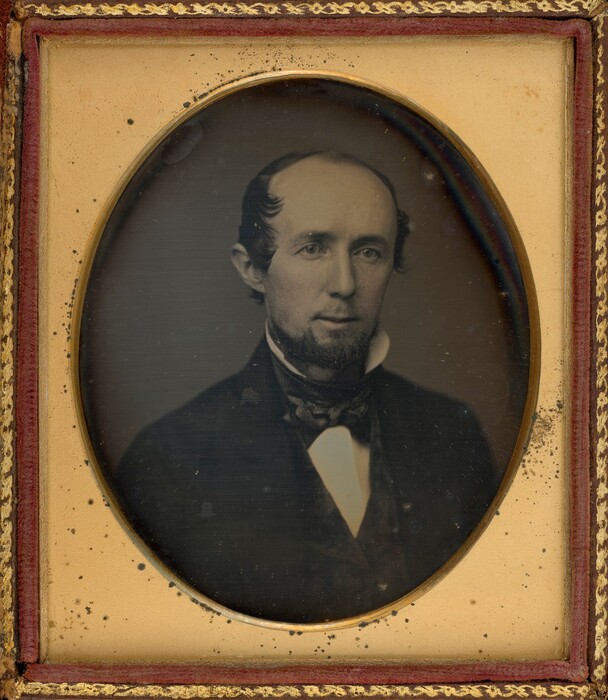 The image shows a head-and-shoulders portrait of a man facing slightly to the right, with his gaze directed towards the camera. His facial features include a well-groomed mustache and beard combination that frames his lips. His hair is combed back from a high forehead, with visible hair receding at the temples but styled to show soft waves. He is dressed formally in a dark coat with a white shirt underneath, along with a cravat or bow tie wrapped around his neck. The photograph is housed in an ornate frame with a gold border and has a plain dark background. The daguerreotype is enclosed in a case with a red velvet lining around the inner perimeter under the glass cover.