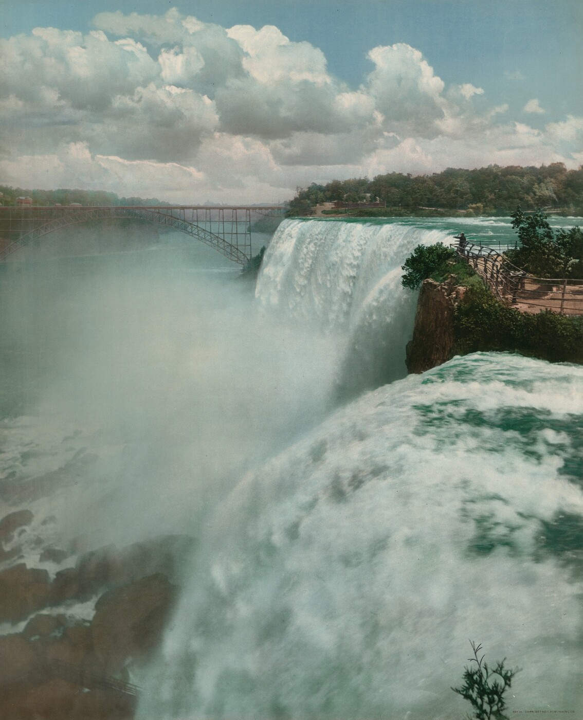 This photograph shows a white-and-blue waterfall on the right cascading over a rocky edge into a misty river below on the left. There are large brown rocks in the bottom left corner, the white spray from the waterfall partially obscuring them. Beside the waterfall, on the far right, is a viewing platform with a fence and greenery. A bridge stretches across the scene in the distance on the left, connecting the land on either side of the pale blue river. Greenery extends into the distance beyond the bridge. The sky has fluffy white and gray clouds against a pale blue background.