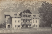The image shows a large, ornate building with multiple windows and balconies. A wooden picket fence is visible in the foreground, with grass below. In the background, there are majestic mountains with snow-capped peaks. A tree stands to the right of the building. The scene is in sepia tones, giving it a vintage feel.