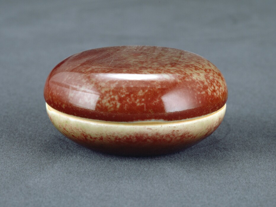 The sculpture is a round, smooth container with a lid. It is made of glossy ceramic material in a rich, reddish-brown hue with a speckled pattern. The bottom part is a lighter beige with a speckled appearance. The sculpture has a polished look, smooth edges, and a reflective surface. It appears compact, finely crafted, and showcases refined artistry and craftsmanship.