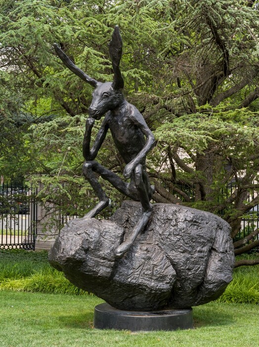 A rabbit with long ears and spindly limbs sitting on a boulder is cast in bronze in this freestanding sculpture. In this view, the rabbit’s body is angled to our left. It rests its long muzzle on its right fist, to our left. That elbow rests on the right, bent knee. The other wrist rests on the other bent knee. The textured boulder sits on a disk-like round base also cast in bronze, and the sculpture is displayed on a grassy lawn against a screen of mature green trees.