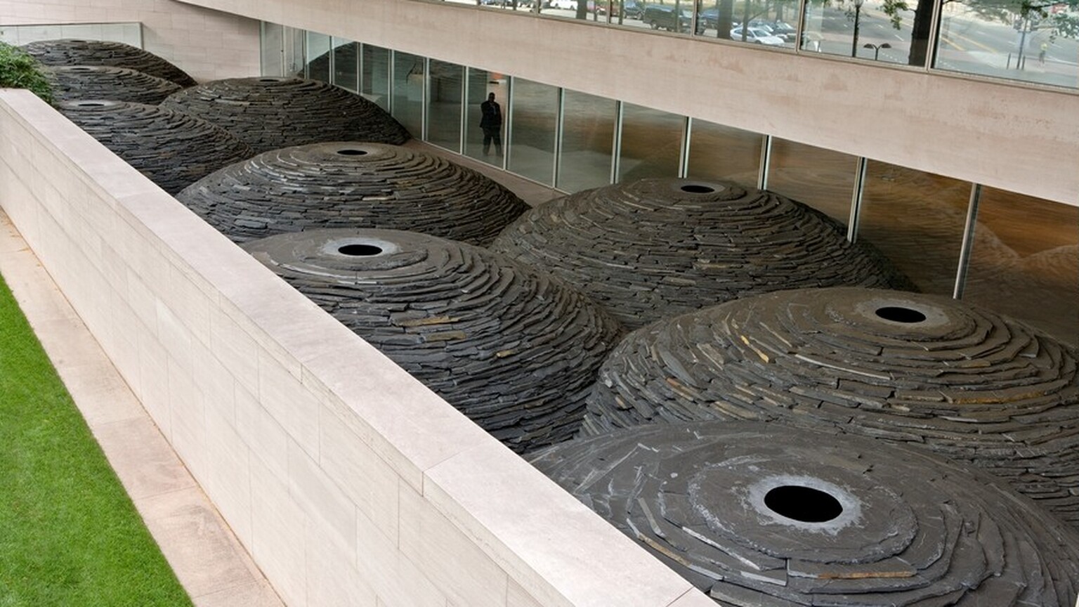 We look down onto nine gray stone domes that melt into each other in the space of a patio enclosed with a pink marble wall opposite floor-to-ceiling windows. Each dome is nearly as tall as the ceiling over the windows, and each is made of stacked pieces of slate. At the top center of each dome is a collar of gray slate to create a pupil-like opening. The stone collar creating the openings is lighter gray around the ink-black centers, which are the shaded interiors of the domes. The domes meld and stack like a handful of bubbles.