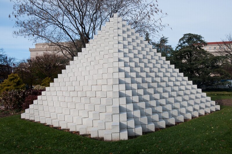 A free-standing, stepped pyramid made of interlocking, white concrete blocks sits on a grassy lawn, rising above the bushes and low trees behind it. This photograph shows the pyramid with one corner coming straight at us so each side is a zigzag of twenty-four steps. The structure is lit from our left so casts gray shadows to our right. One tall tree behind the pyramid extends off the top edge of the photograph and buildings in the distance have cream-white walls and red roofs.