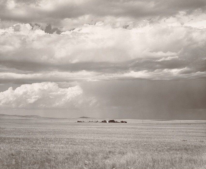 The image shows a wide, expansive landscape with a vast, open field extending toward the horizon. Sparse clouds cover the sky, casting a soft light over the scene. The land is covered with dry grass, leading to a cluster of structures in the distance. The photo is monochromatic, using a range of grays to highlight texture and tone.