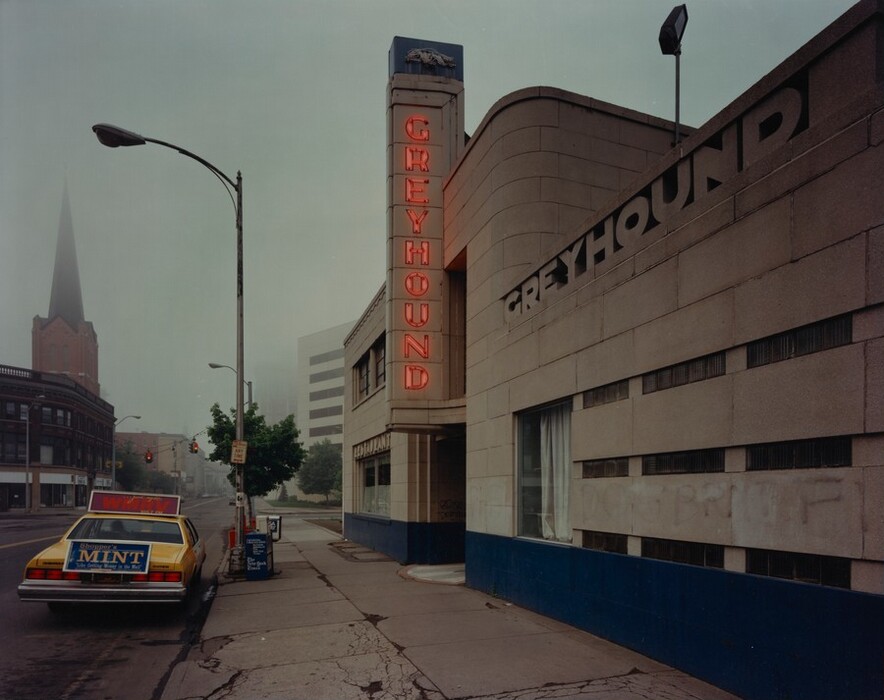 The image shows a street-level view of an urban scene with a Greyhound bus station on the right side of the frame. Tall buildings and slight fog obscure the horizon. The Greyhound sign is illuminated in red on the concrete building exterior. A yellow car is parked on the left side with advertisements. A streetlamp and traffic lights line the street heading towards a brick building with a tall spire in the distance. The color palette is subdued with grays and earth tones, highlighted by the red signage.