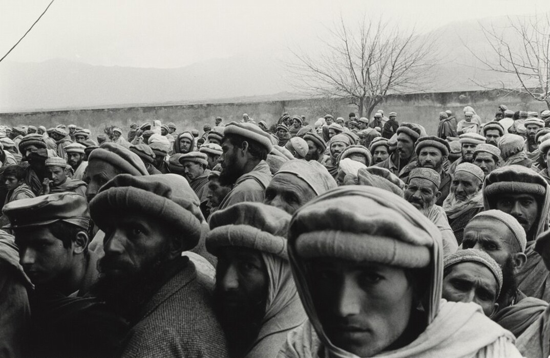 The image shows a large crowd of men standing close together outdoors. They have diverse facial features, with most of them having mustaches and beards. Many are wearing traditional caps like pakol, and some have warm clothing such as jackets and shawls. One man wears a distinctive leather cap. The background includes a bare tree, a low wall, and misty, mountainous terrain. The photo is in black and white.