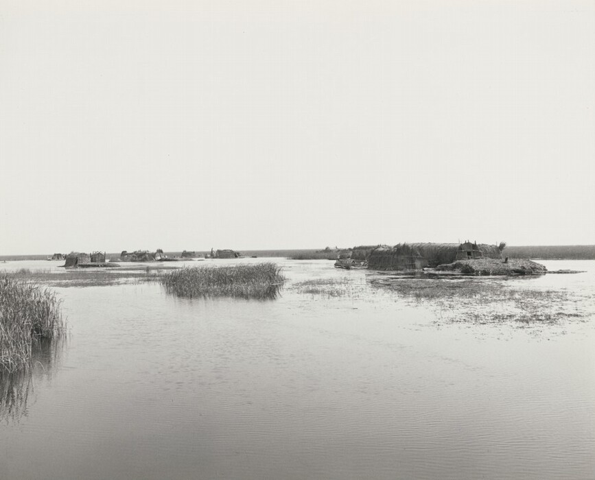 The image shows a body of water with traditional huts in the distance. Near the viewer, there are patches of reeds and aquatic plants. The color palette is monochromatic with varying shades of grey.