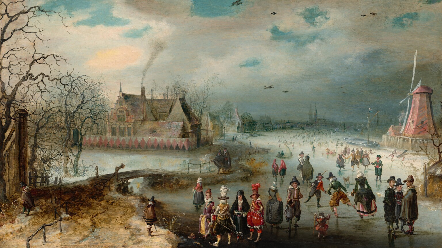 We look slightly down onto a scene showing light-skinned men, women, and children ice skating on a frozen river in this horizontal landscape painting. We get the impression of hundreds of people gathered on the ice creating a crowd that extends into the hazy distance. The couple dozen closest to us are the most defined. A few clusters of people and individuals in particular draw our attention. For example, a group of three men wearing dark cloaks and hats stand in conversation on our right. Two boys nearby hold sticks and play a game similar to hockey. A small child holds two smaller sticks, perhaps to help balance. A man in the front center wears puffy scarlet-red pants with white stockings, a red jacket, and a tall brown hat with a cloud of scarlet feathers. He stands next to a woman wearing a black hooded cloak over a black skirt and raspberry-pink bodice. She tucks her hands into a cylindrical muffler held at her waist. Another elegantly dressed man in golden yellows and black and a woman in mauve pink and butter yellow stand nearby. Some of people throughout the scene wear white frilly collars and others are more simply dressed in shades of brown, gray, and black. To our right, a faded rose-red windmill stands at the river’s edge, and to our left a large house with steeply pitched and stepped roofs is enclosed within a solid fence painted with pink diamonds against black and white. Smoke rises from one chimney, and other houses and a church line the riverbank into the distance. A wooden bridge near a grove of bare trees connects spit of land near us in the lower left corner with the village beyond. Brilliant azure-blue sky is visible through breaks in the steel-gray clouds above.