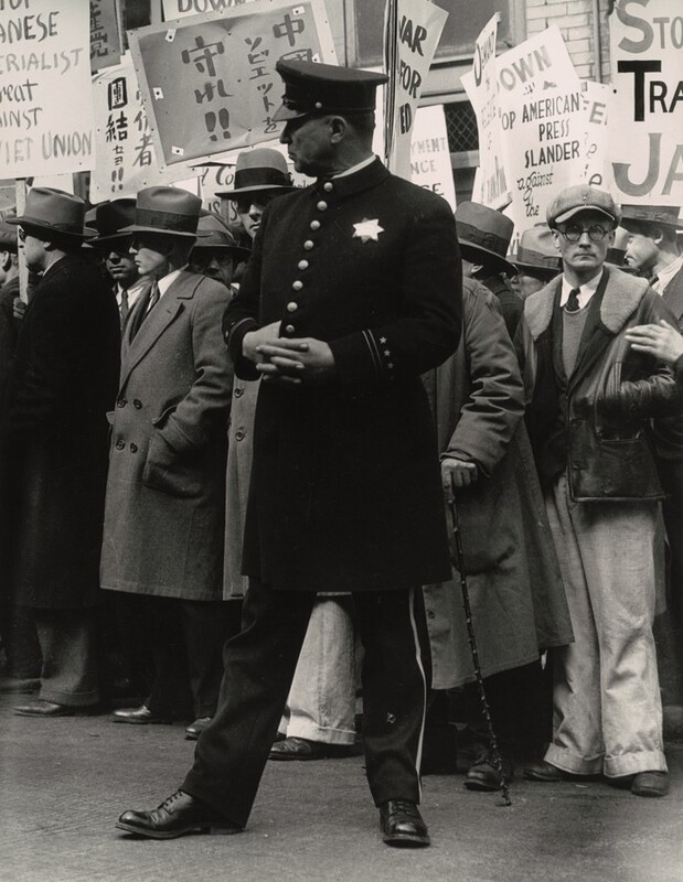 A uniformed policeman standing in front of a dense crowd of men holding protest signs in English and Japanese nearly fills the height of this vertical black and white photograph. The policeman either has a ruddy complexion or his face might be in shadow. He stands with his body facing us but he turns his head to our left in profile. His hands are crossed over his chest and one thumb is hooked into his jacket between two of the eight brass buttons down the front. A seven-pointed star reflects light on his chest and his flat-topped hat has a short, shiny brim angled down over his eyes. The jacket comes to his knees and his pants are lined with gold or other light material down the side. His feet are widely planted, and he wears dark shoes. The men in the crowd behind appear to be light skinned. Many wear button-down, collared shirts with ties tucked into long coats. Many also wear fedora hats and look off to our left, the same direction as the policeman. One man to our right wearing a floppy cap and glasses looks out at us with his hands in his jacket pockets. Some of the legible English words on the protest signs read “UNION,” “AMERICAN PRESS SLANDER against the,” and “JA.”
