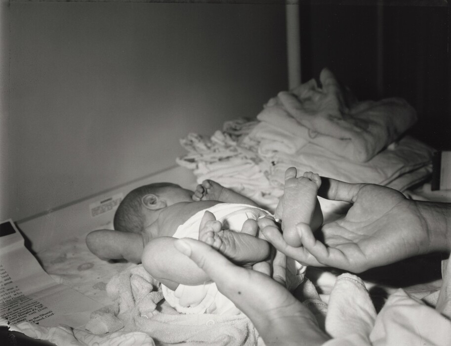 The image displays a newborn baby lying on its back. The baby's head is turned to the side, partially obscuring the face. The baby is wearing a diaper and is placed on a soft surface, likely a changing table, with a textured blanket underneath. An adult's hands are holding the baby's feet. In the background, there are neatly folded stacks of fabric or clothing. A piece of paper with text on it is visible near the baby. The setting seems to be indoors, possibly in a nursery or hospital.