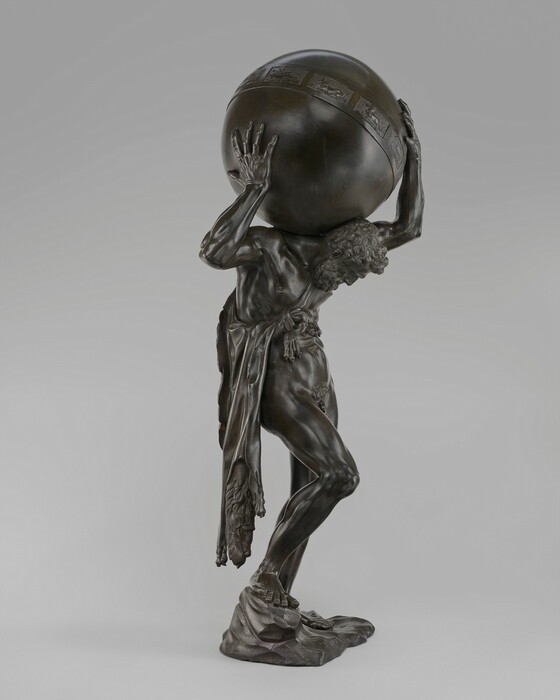 The sculpture depicts a muscular male figure in a bent-over position, supporting a large globe on his shoulders. The figure is nude, emphasizing the detailed musculature and physical strain of holding the globe, which is textured with decorative bands. The sculpture is made from a dark bronze material. The man is depicted in a dynamic pose, with one knee slightly bent and the other leg forward. The sculpture stands on a rocky base.