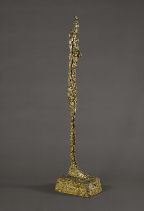 An elongated, abstracted nude woman stands with her arms pressed to her sides on oversized feet on a blocky plinth in this free-standing bronze sculpture. The woman is unnaturally thin and has shoulder-length hair and low-set breasts. The rough, knubby surface of the bronze is gold colored. In this photograph, the woman is angled to our right, and she is shown against a charcoal-gray background.