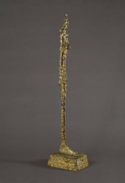 An elongated, abstracted nude woman stands with her arms pressed to her sides on oversized feet on a blocky plinth in this free-standing bronze sculpture. The woman is unnaturally thin and has shoulder-length hair and low-set breasts. The rough, knubby surface of the bronze is gold colored. In this photograph, the woman is angled to our right, and she is shown against a charcoal-gray background.