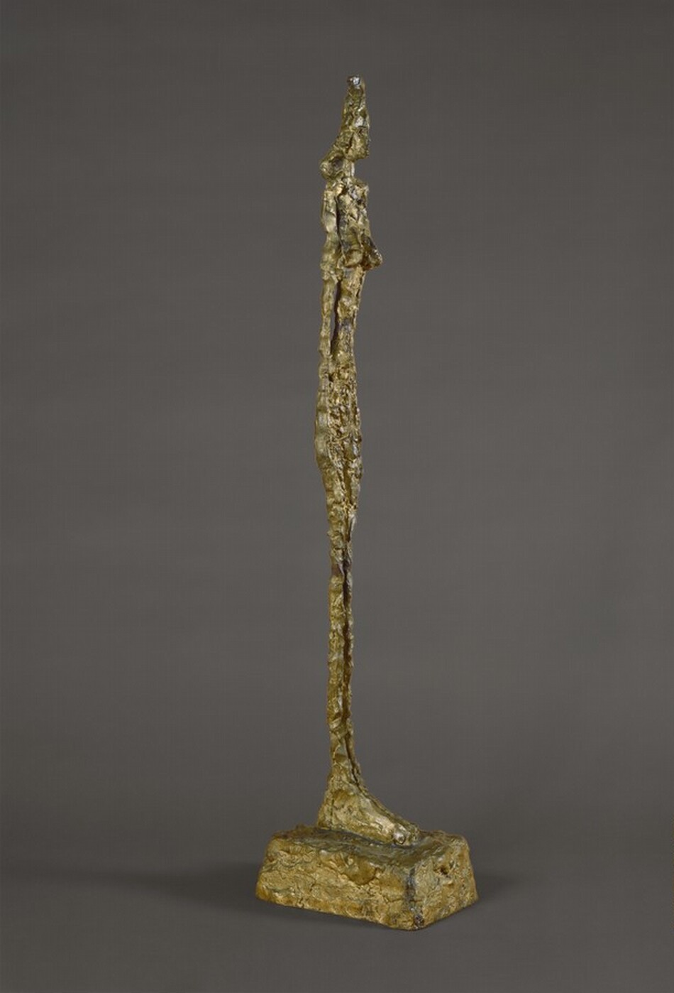 An elongated, abstracted nude woman stands with her arms pressed to her sides on oversized feet on a blocky plinth in this free-standing bronze sculpture. The woman is unnaturally thin and has shoulder-length hair and low-set breasts. The rough, knubby surface of the bronze is gold colored. In this photograph, the woman is angled to our right, and she is shown against a charcoal-gray background.